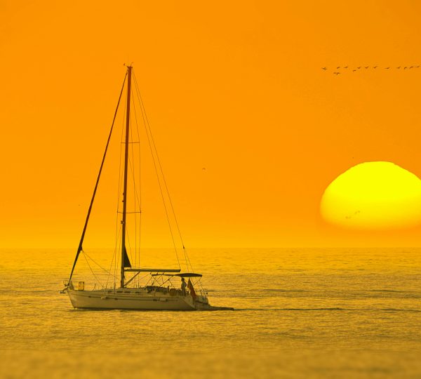 A luxury yacht sailing smoothly at sunset in the Strait of Gibraltar, with the coast of Tangier or the outline of the Kasbah visible in the distance and a sky filled with warm colors. Enjoy your Luxury Sailing in Tangier.